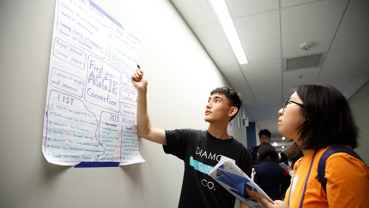 Two students stand in a hallway and look at a white poster on the wall with handwritten notes titled "First Year Aggie Connections." One student is speaking and points to the poster as the other student holds and open notebook taking notes while listening and looking at the poster.