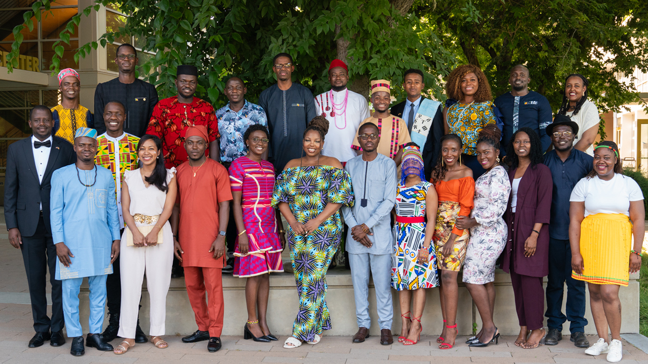A cohort of 25 Mandela Washington Fellows stands under a large tree outside in colorful traditional attire.