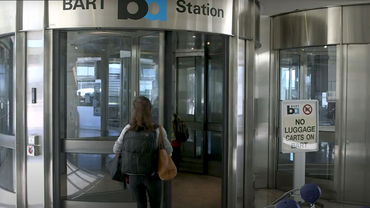A woman with bags walk through a revolving door marked into a BART station.