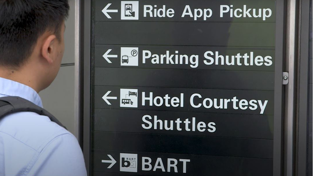 A man looks at a black sign with arrows directing where to find Ride App Pickup, Parking Shuttles, Hotel Courtesy Shuttles and BART.