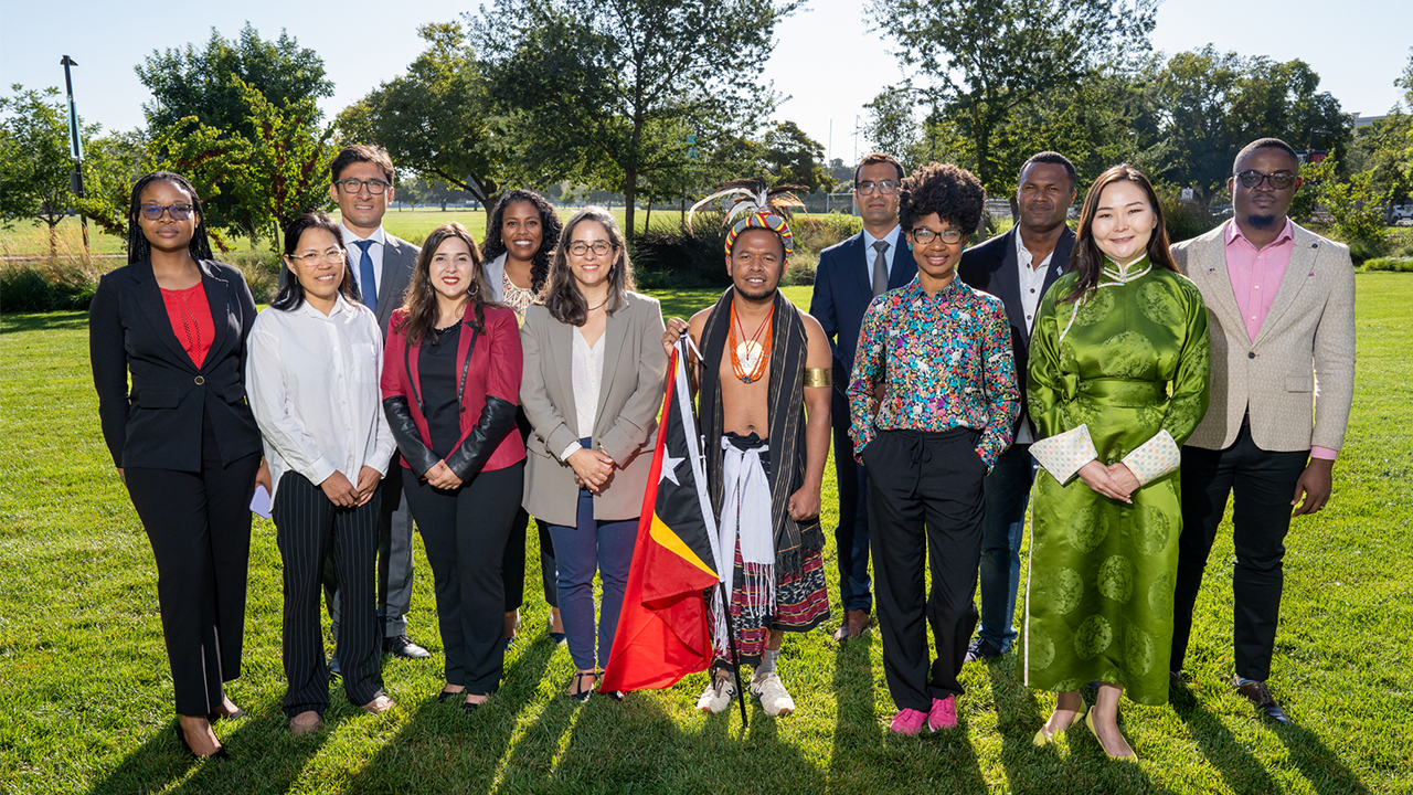 Twelve Humphrey Fellows stand together in two rows on the grass of a lawn on the UC Davis campus in the sunlight. They wear a combination of culturally traditional and business dress. 
