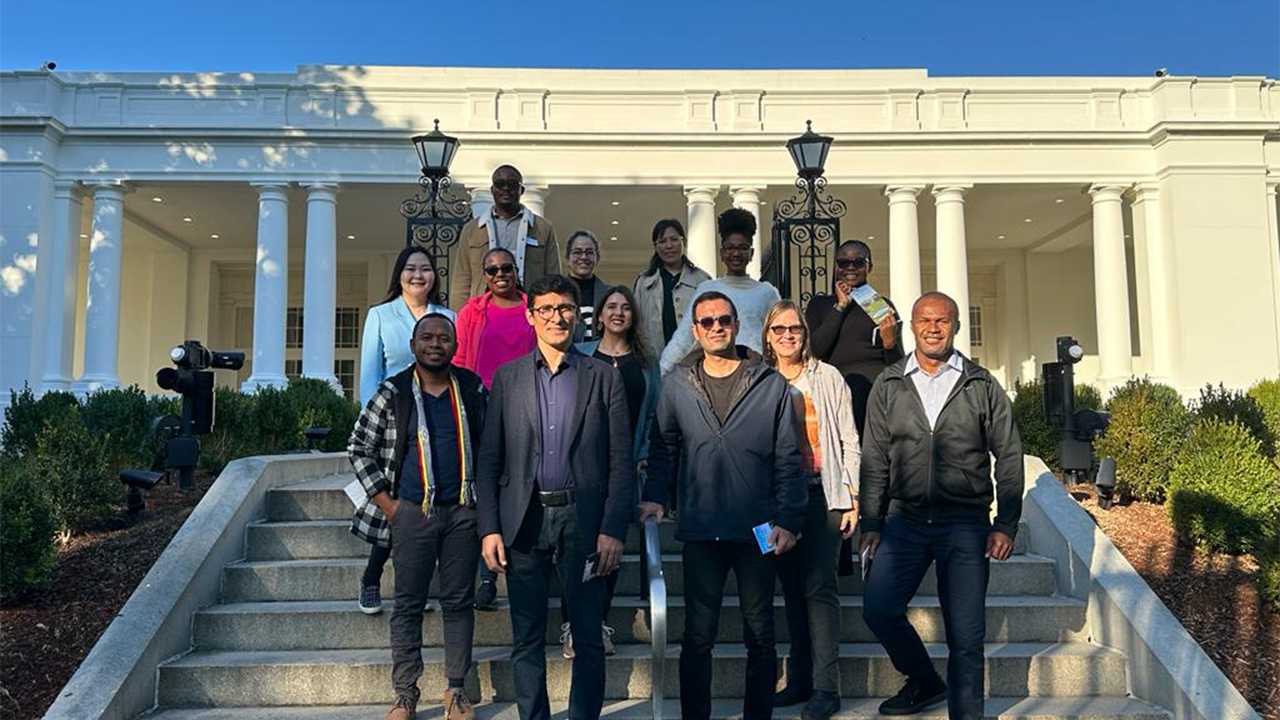 The Fellows stand on steps in front of the White House that lead up to the white columns and open breezeways behind them.