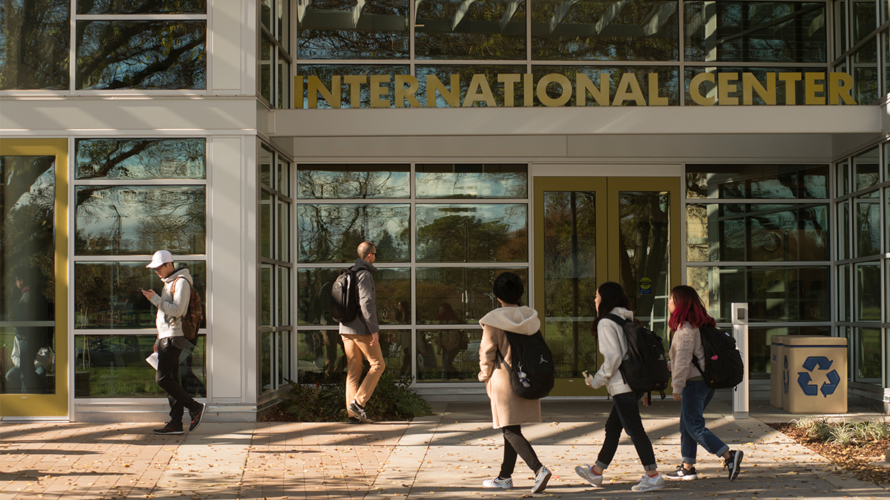 On a sunny day, three students dressed for cool weather and wearing backpacks approach the doors of the International Center on the right side of the photo. Another student wearing a jacket and backpack approaches from the left, and another students looks at their phone while walking away from the building at the very left of the photo.