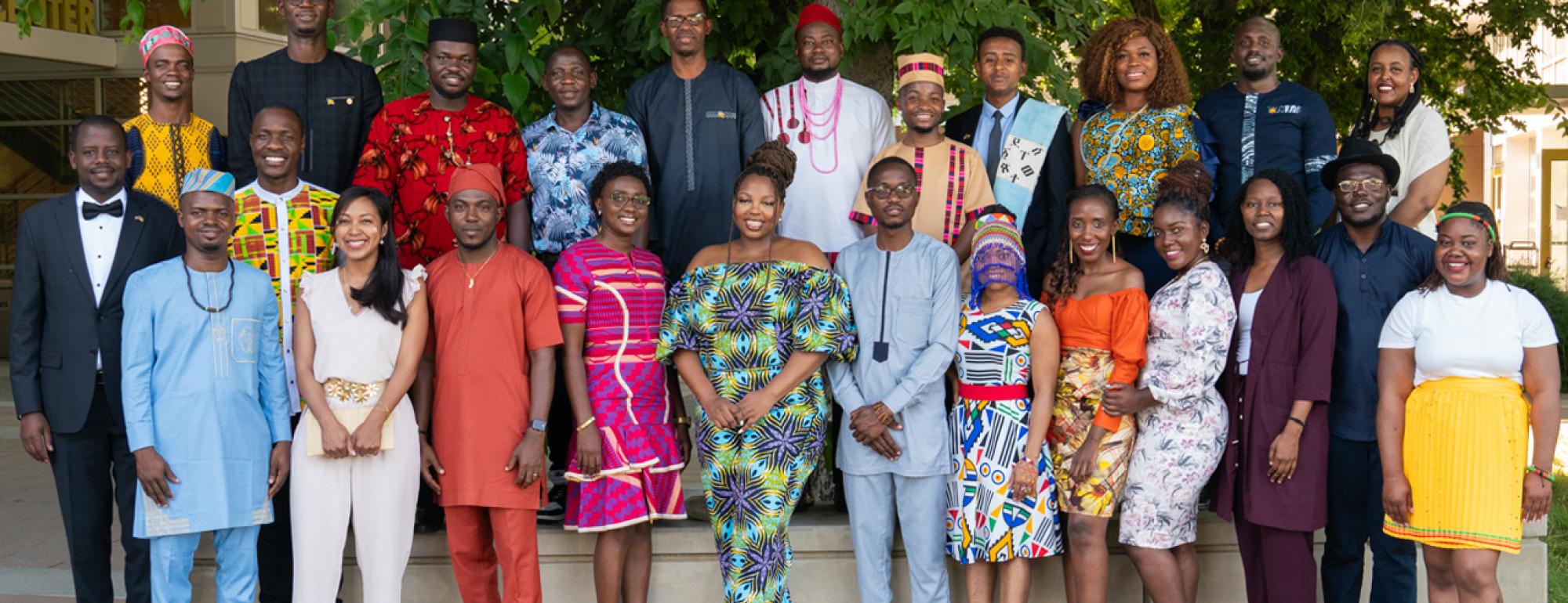 A cohort of 25 Mandela Washington Fellows stands under a large tree outside in colorful traditional attire.