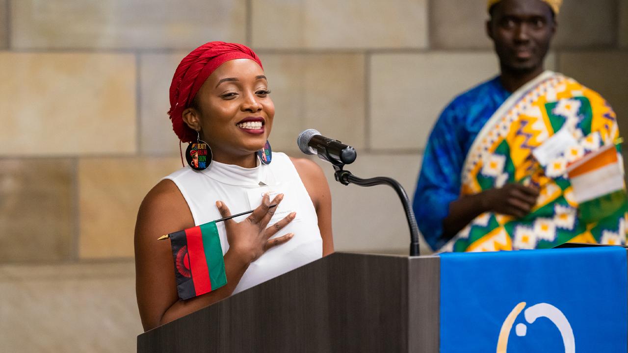 Melissa stands at a podium. She wears a red scarf wrapped around her head, a sleeveless white top with a high collar and earrings with the phrase "Beauty Has No Skin Tone". She hold a Malawian flag in her right hand and holds the hand to her heart.