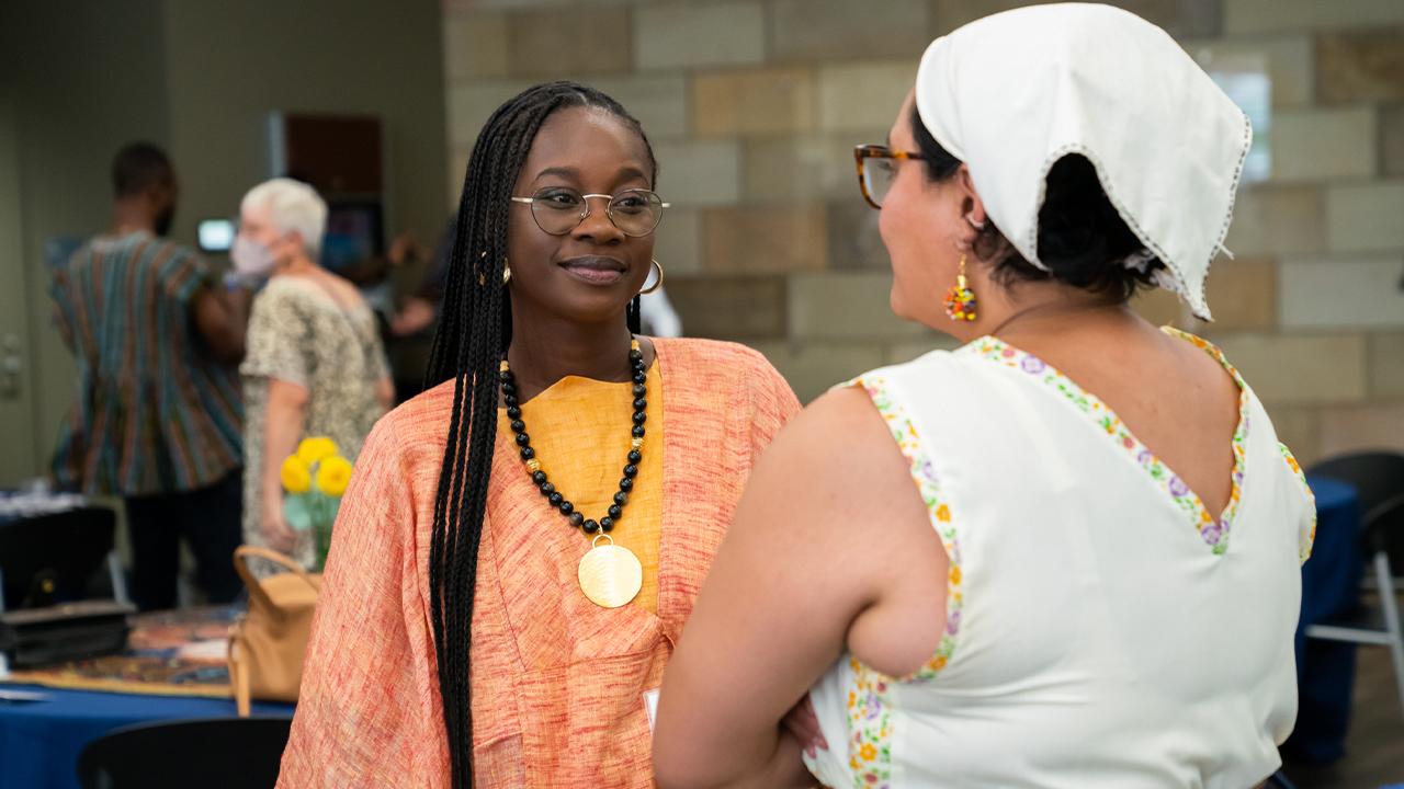 Yaye wears an orange and yellow linen tunic with a black beaded necklace with a gold medallion. Her earrings are gold hoops and she wears round metal glasses. She speaks with a woman whose back is to us, but who wears a scarf on her head and a white blouse that dips to a V in the back.