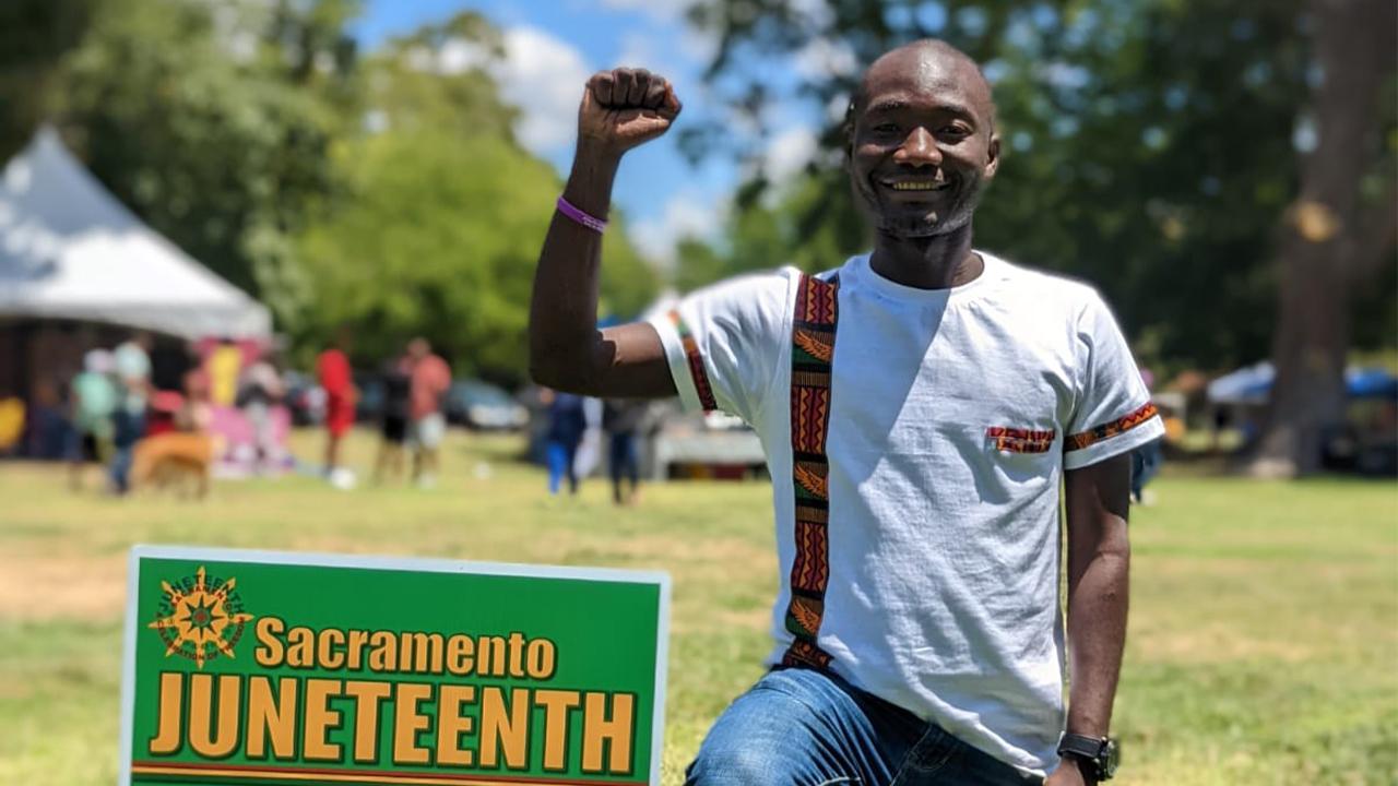 Outside in the sunlight, Zengani kneels on his left knee next to a green sign with yellow writing that says Sacramento Juneteenth. His right fist is raised in solidarity with the African American community. He wears blue jeans and a white T-shirt with embellishments in tribal patterns in red, orange, green and black. Behind him, people gather by trees and a white tent.