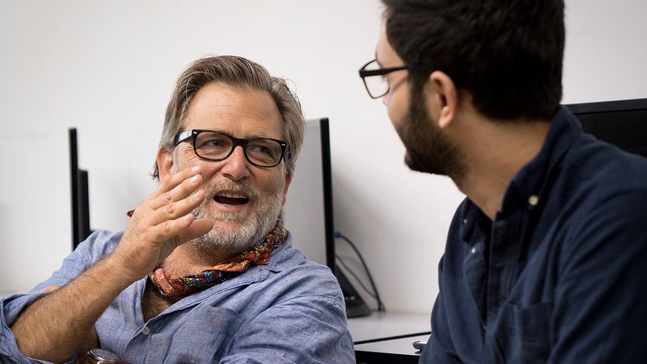 Keith Watenpaugh (left) in a chambray blue collared shirt, speaks to a student on the right. Keith has salt and pepper hair, a gray beard and wears black rectangular framed glasses. The student is turned so we can't see his face, but we can see he has dark short hair, a dark beard, wire framed glasses and wears a navy collared shirt.