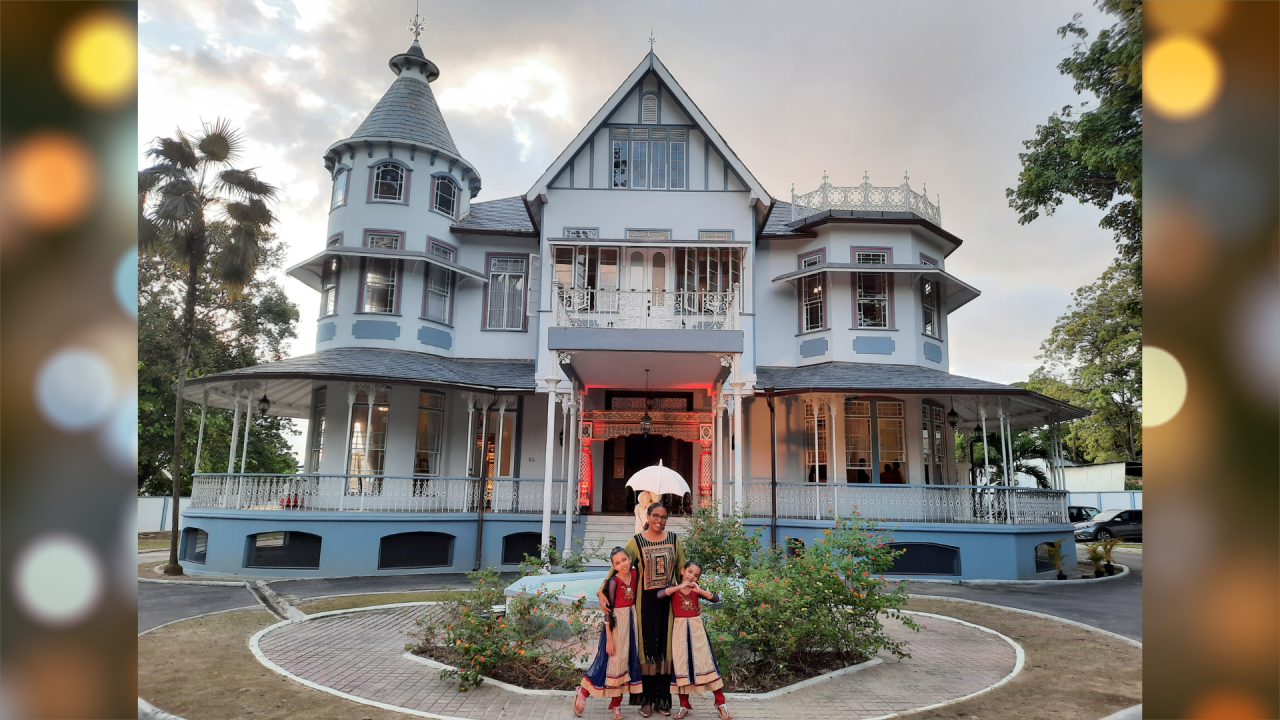 Kara Roopsingh stands in front of a light blue two-story wooden French provincial-style house with a wrap-around porch and frilly cast iron railings. With Kara are two little girls dressed identically and smiling with her. 
