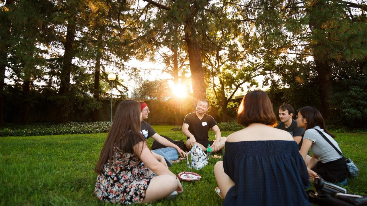 Students sitting around in a circle on the lawn. There is a sunset in the background.