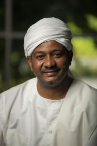 Ahmed Osman Elamin Abdalgader smiles in this 2021 headshot. He has a short mustache and wears a white turban and white traditional Sudanese dress.