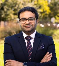 Ahsan stands outside with his arms crossed casually and his large round smart watch face visible on his left wrist. He wears dark rimmed rectangular glasses, a navy suit, white shirt and navy tie with thin white and red stripes.  tie