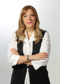 Lina poses with her arms crossed in a white collared shirt with a black vest in front of a white background.