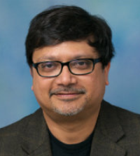 Sudipta poses in a gray suit and a black shirt in front of a blue background wearing glasses.