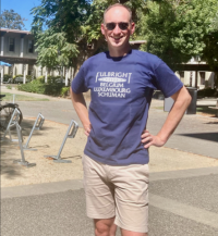 Tom smiles with his hands on his hips in a blue Fulbright t-shirt and khaki shorts with the U.C. Davis campus in the background.