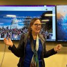 Karen stands at the front a a room with two large monitors behind her displaying various interfaces. Her hands are raised, palms up, nearly shoulder height as she speaks.