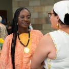 Yaye wears an orange and yellow linen tunic with a black beaded necklace with a gold medallion. Her earrings are gold hoops and she wears round metal glasses. She speaks with a woman whose back is to us, but who wears a scarf on her head and a white blouse that dips to a V in the back.