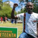 Outside in the sunlight, Zengani kneels on his left knee next to a green sign with yellow writing that says Sacramento Juneteenth. His right fist is raised in solidarity with the African American community. He wears blue jeans and a white T-shirt with embellishments in tribal patterns in red, orange, green and black. Behind him, people gather by trees and a white tent.