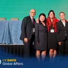 Bob Kiaii stands in front of a stage with three woman and one man all wearing business attire. The stage behind them is set up for a panel of speakers with draped tables and chairs in front of a large light green screen. A graphic with the UC Davis Global Affairs logo and #GlobalAggies lays over the photo at the bottom.