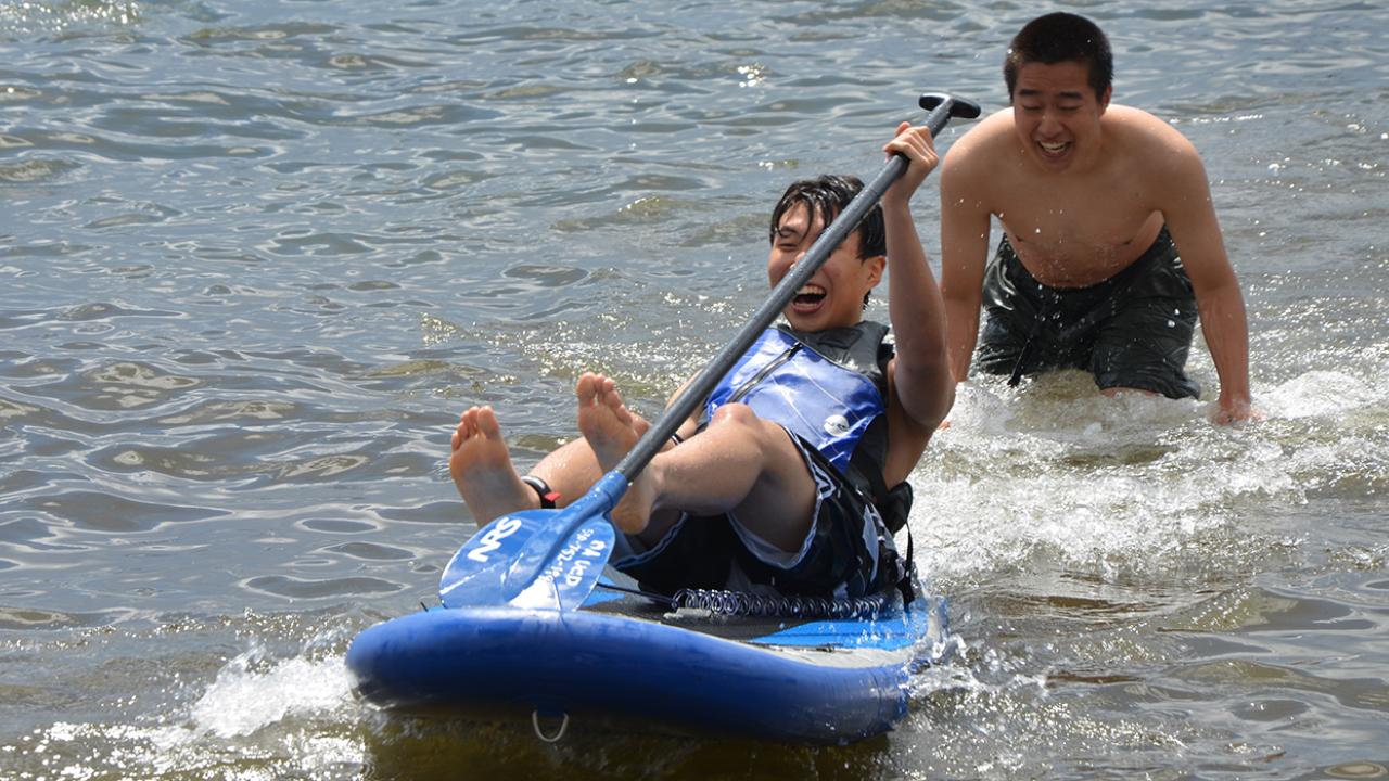 Two students play in lake water. One in a life jacket smiles joyfully and sits leaning back slightly on a blue paddle board with an ore as it propelled forward. The other stands thigh deep in the water bent over in action laughing at the first student. The water splashing around them suggests the student standing launched the student on the board.