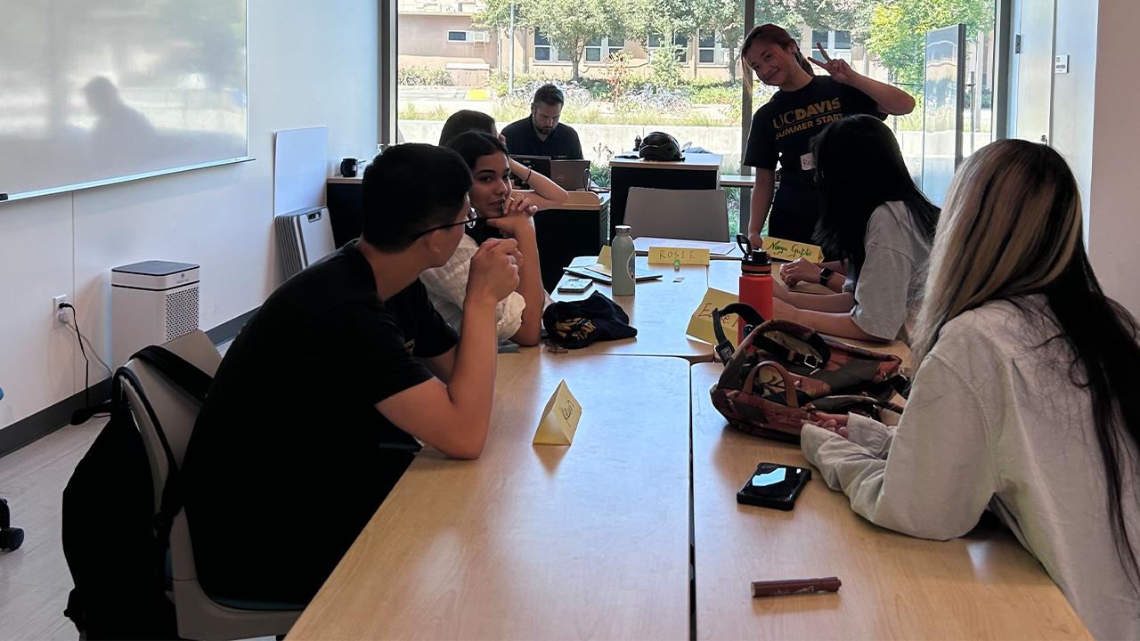 Six students around a table with their instructor working on a laptop at the front of the room. One student in a UC Davis T shirt stands and gives a peace sign with two fingers to the camera.