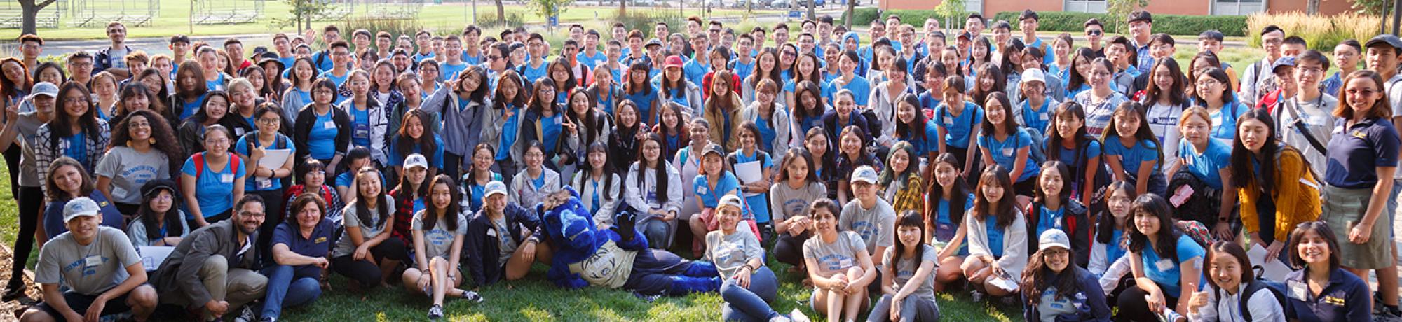 A group photo of close to 200 students gathered in a half circle on a lawn surrounded by trees on the sides. 